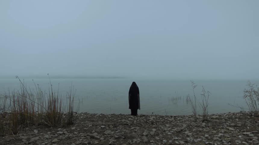 Someone wearing a black cloak over their head and body is standing on the shore looking at the water