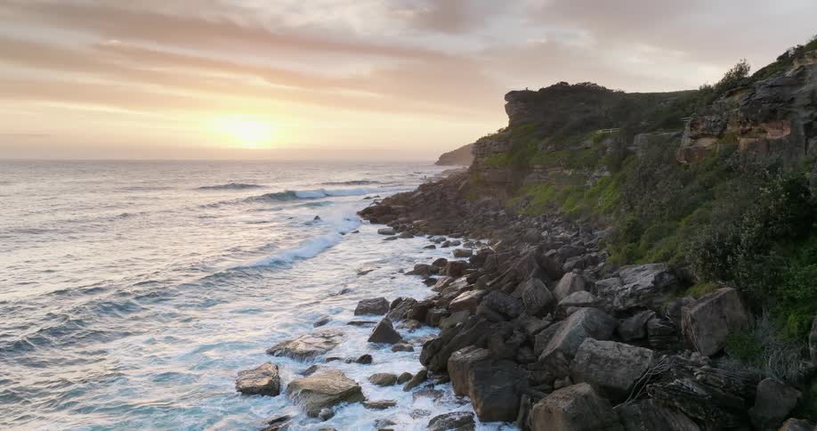 Aerial of gentle waves crashing on a rocky shoreline on the coast at sunrise