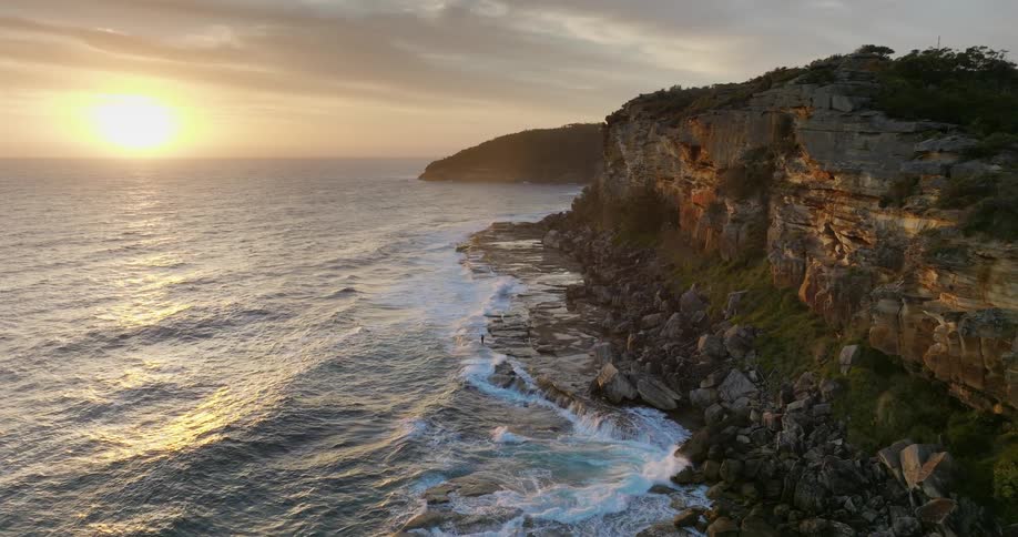 Waves roll over a rocky shoreline as the sun shines on the horizon