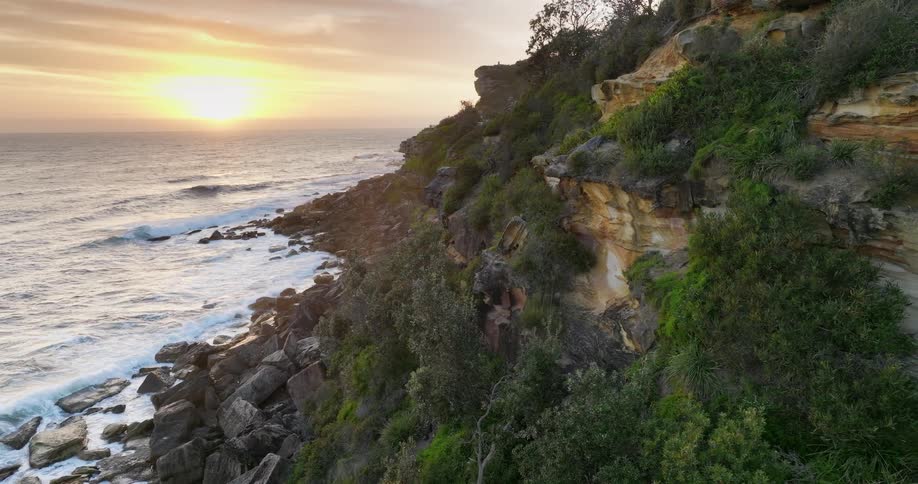 Waves roll against boulders on shore as sun shines from red and gold sky onto promontory