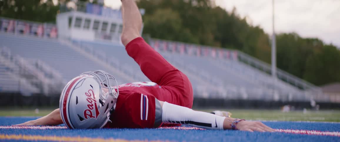 A teenage boy wearing a football uniform is lying on a football field doing leg exercises