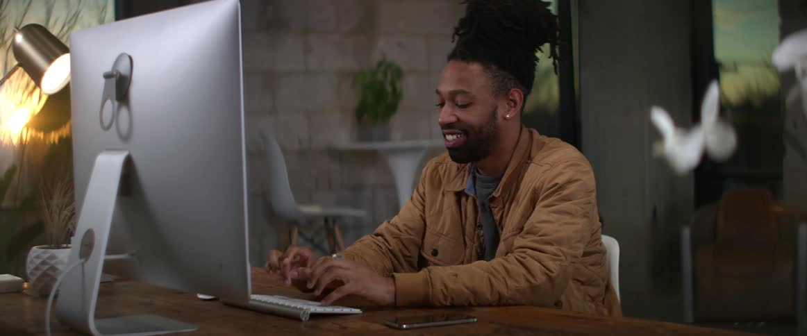 A man is sitting at a desk smiling as he types on a computer keyboard
