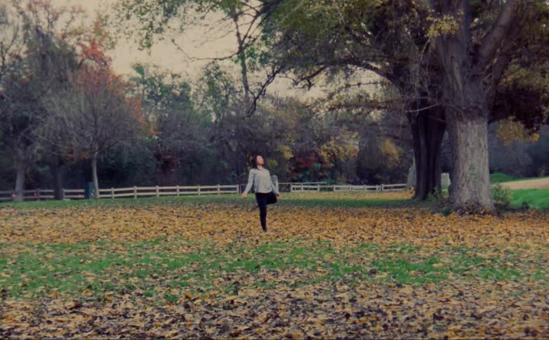 Woman with gray shirt and black leggings dances on lawn with brown leaves in public park