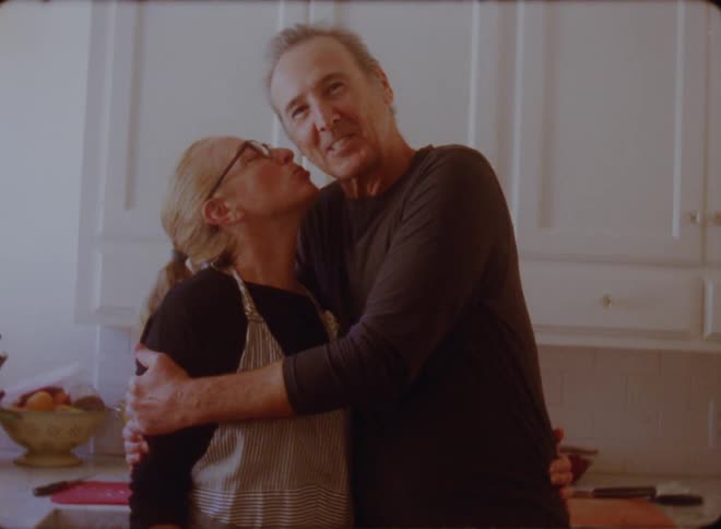 Man with black sweatshirt holds woman with ponytail glasses and apron kissing him in the kitchen
