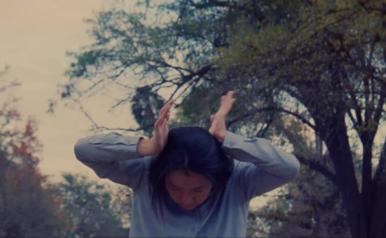 A woman dances with her hands around and on her head in a park