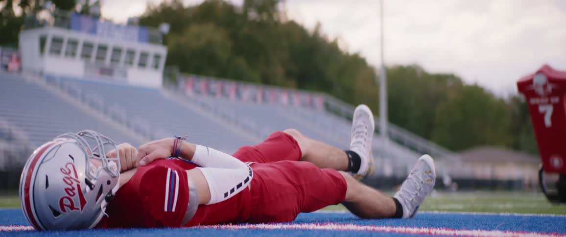 Football player wearing red uniform stretching on the ground on a stadium at evening