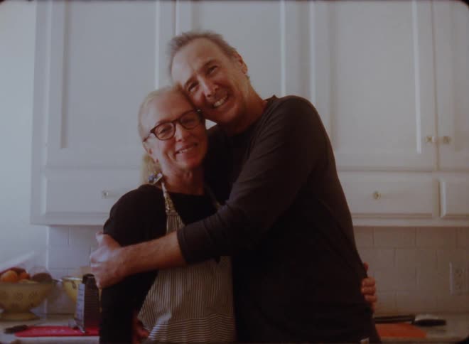 Son with black sweatshirt holds mother with glasses and apron looking at camera in the kitchen