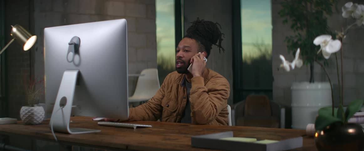 A man talks on the phone while sitting at his desk and using his computer