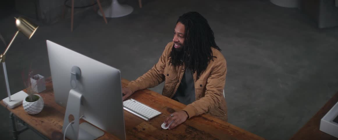 Man with braids and brown jacket sitting at desk types on computer keyboard smiling in office
