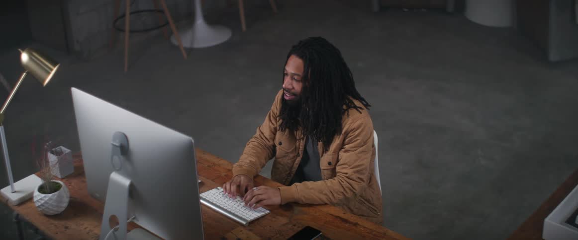 A man smiles as he sits at his desk typing away on a wireless keyboard and looking at the monitor