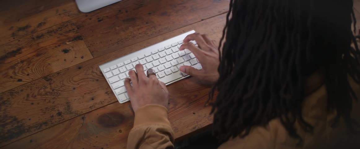 Young man with long curly hair typing on a white computer laptop over a wooden desk