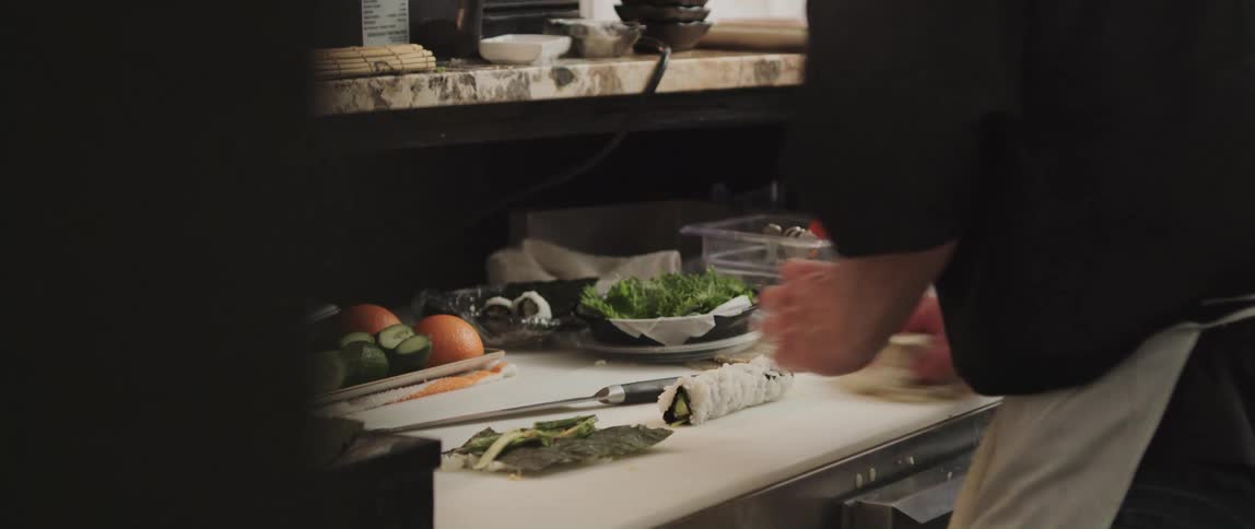 Sushi chef rolls sushi on counter near open cooler