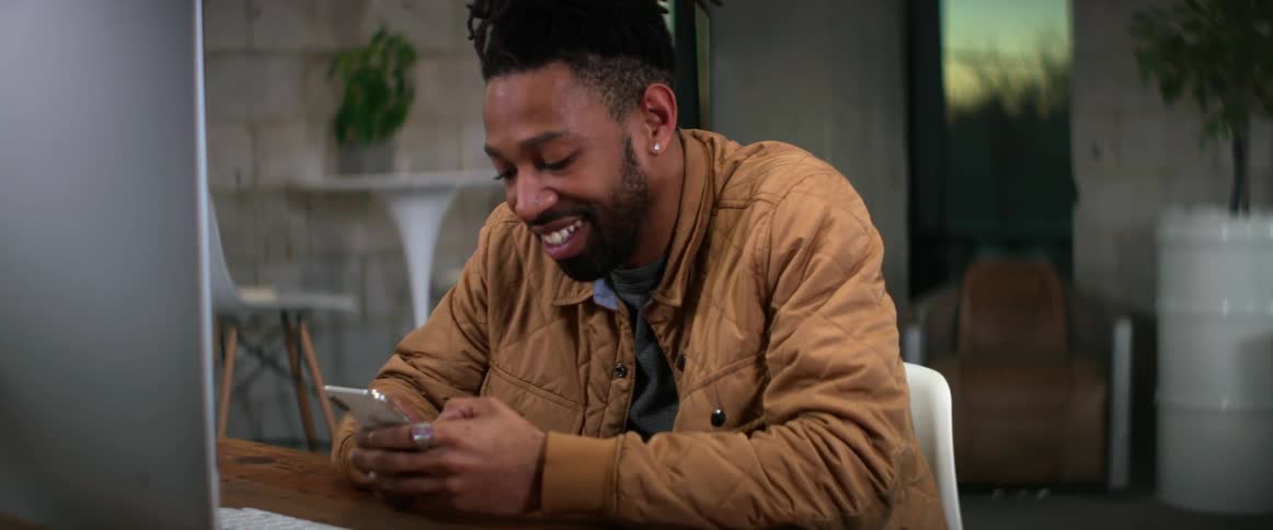 A man smiles while using his smartphone at a desk indoors in the evening