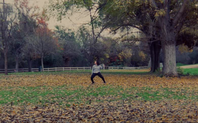 Woman with gray shirt and black leggings dances on lawn with brown leaves in public park