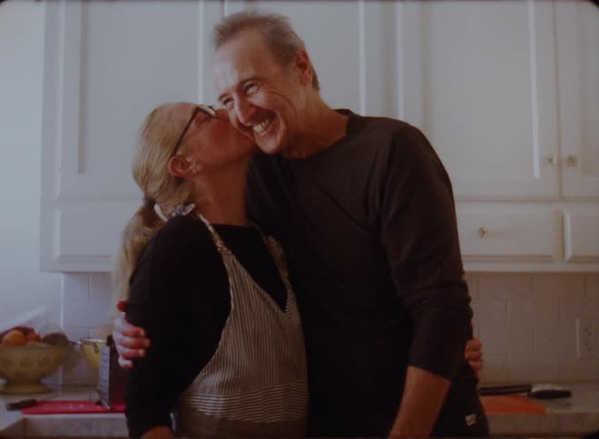 A husband and wife stand in the kitchen and smile and hug and the woman kisses him
