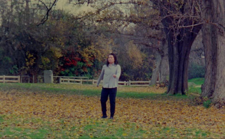 Woman dances in field with woods screening traffic passing in the background