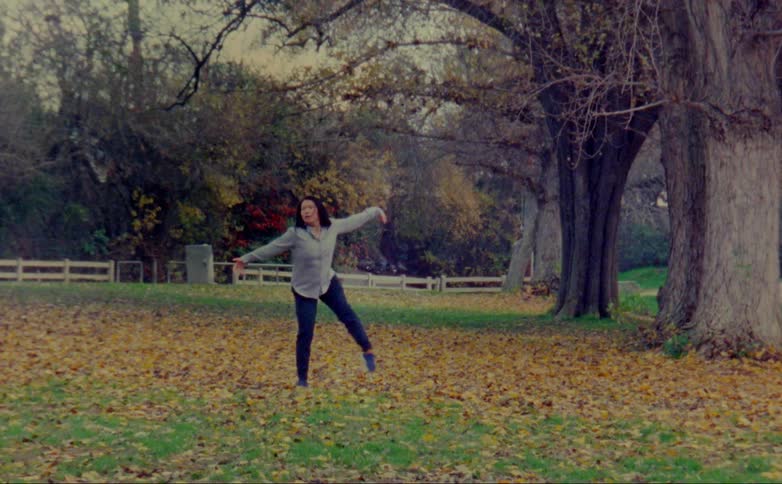 Woman with gray shirt and black leggings dances on lawn with brown leaves in public park