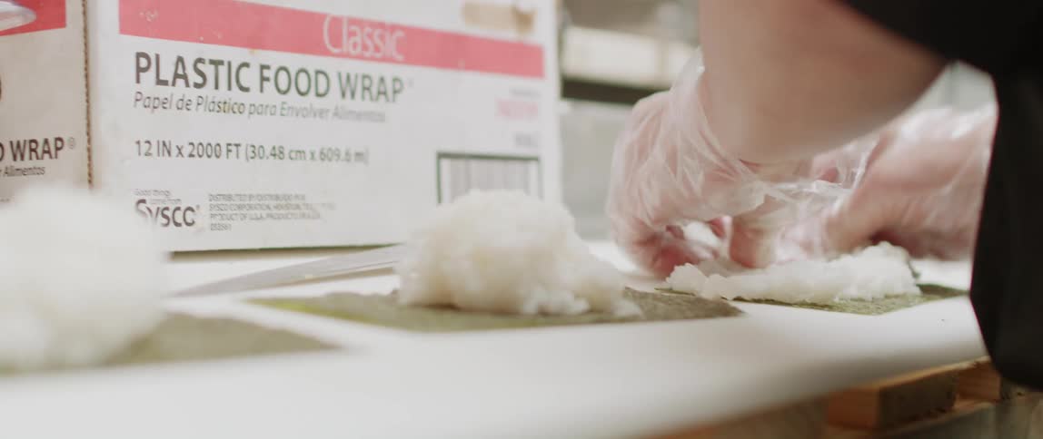 Sushi chef stands at counter near box of food wrap spreading rice onto sushi mat