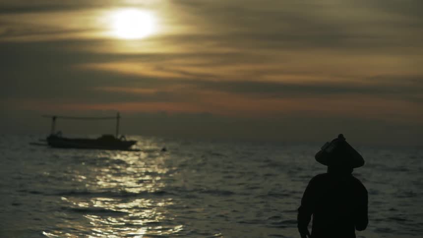 A person is fishing in rough waters with a boat nearby at sunset