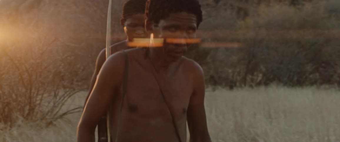 Two indigenous men walk cautiously through grass near a mountain