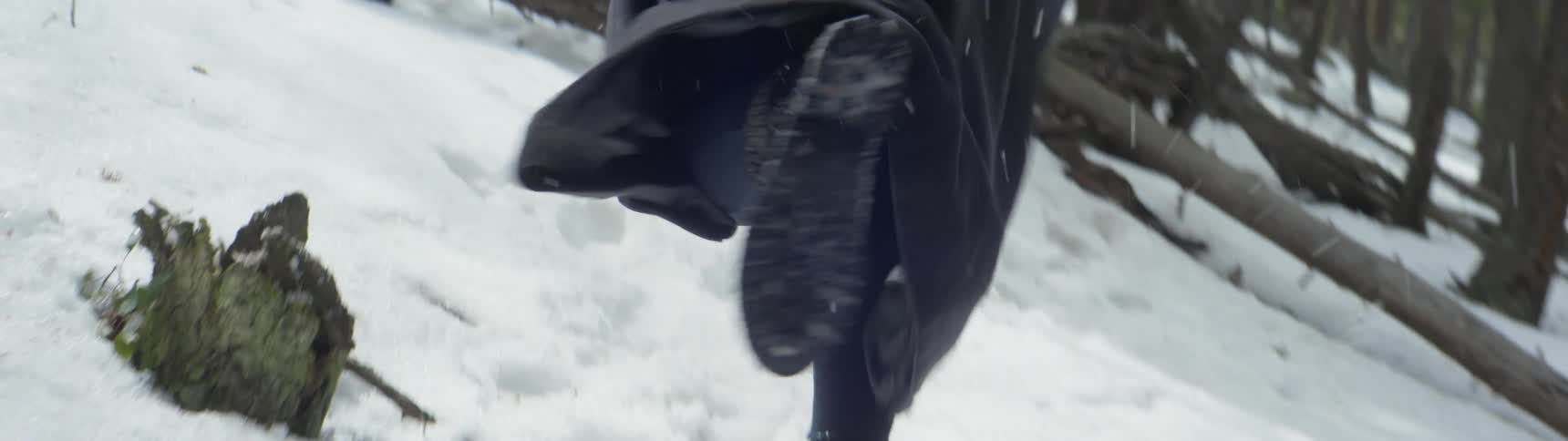 Someone wearing a long dark trench coat and boots is running though a snow covered forest