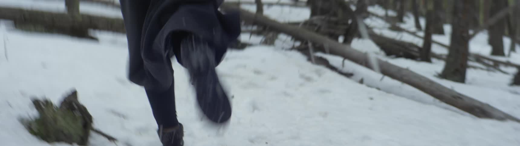 Someone wearing a long dark trench coat and boots is running though a snow covered forest