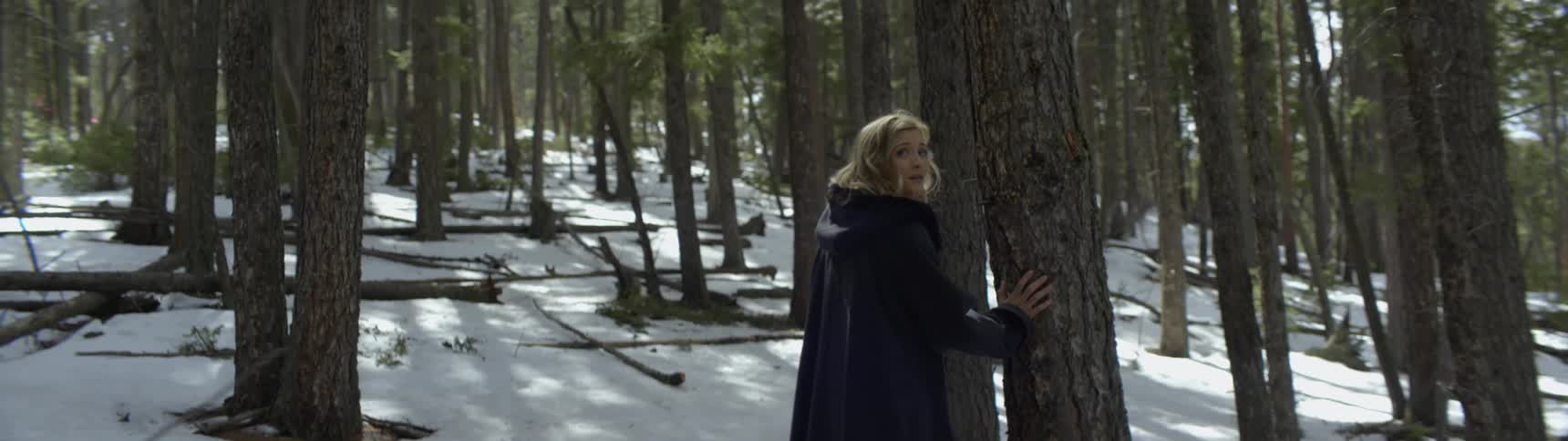 A woman wearing a long dark trench coat and boots is standing near a tree in a snow covered forest