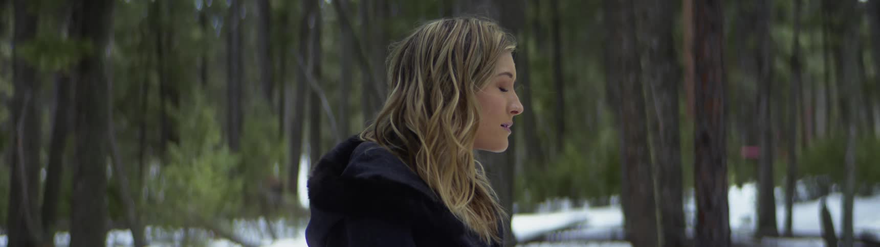 Young blonde woman standing near a forest covered in snow while wind blows her hair