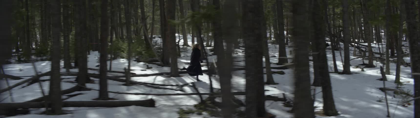 A woman wearing a long dark trench coat and boots is running though a snow covered forest