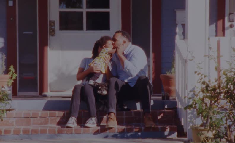 A woman sits on a porch step with her baby daughter standing in her lap and her husband who sits beside her leans over and kisses his daughter