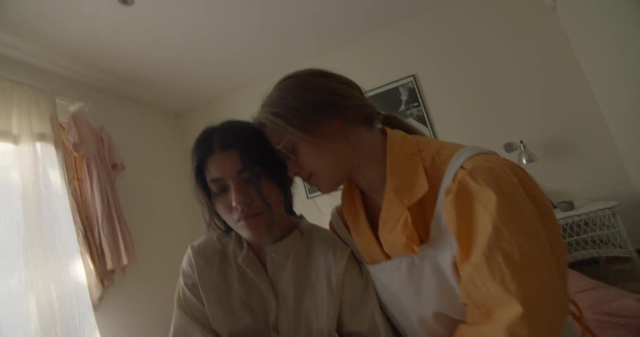Woman with orange shirt and white apron holds woman wearing shirt and skirt kissing and holding hands in bedroom