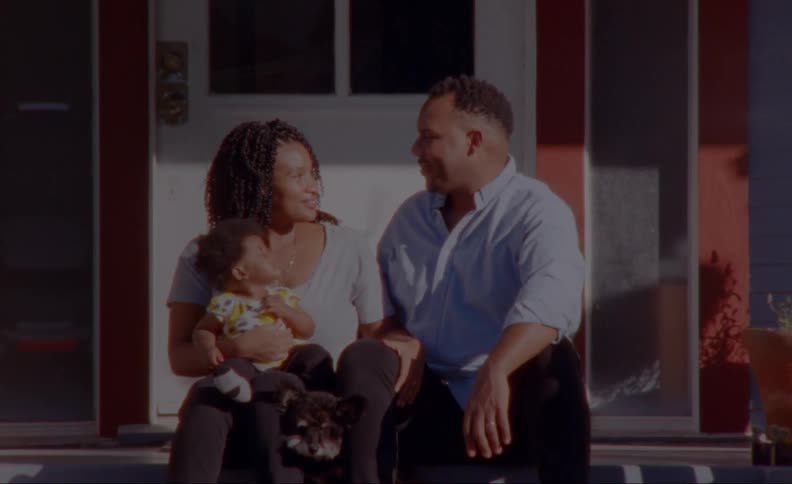 Parents sit on porch with child and pet and smile at each other