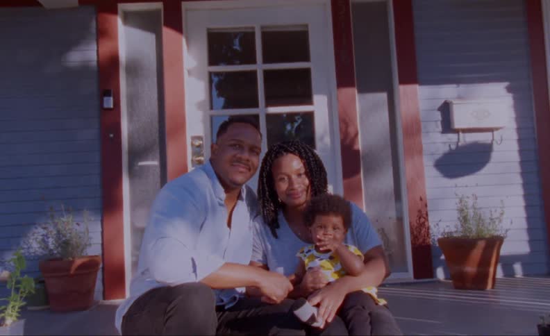 A married couple smile into the camera while sitting on their front step with their young child