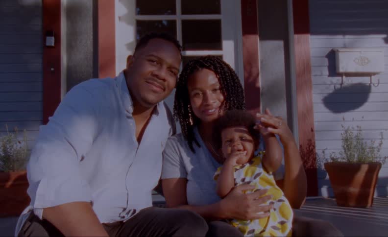 A husband and wife pose on the front steps of a house with their baby daughter