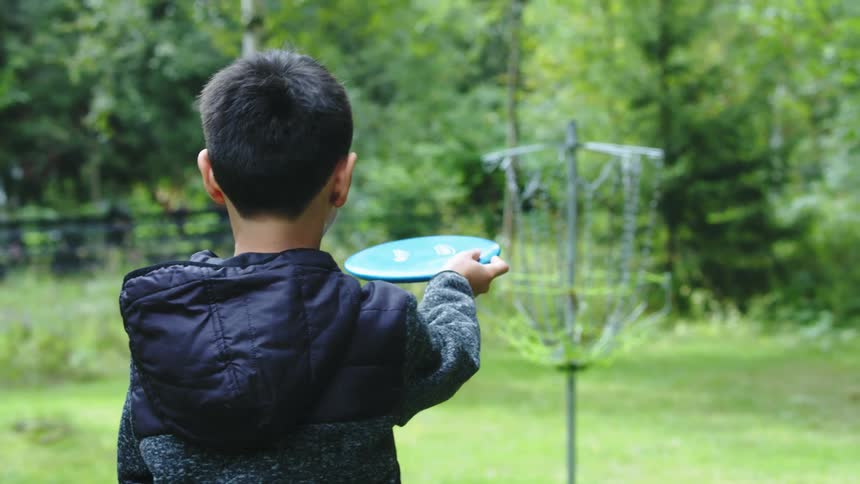 Boy wearing hooded jacket aims disc at target standing in field near woods