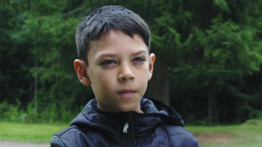 A boy wearing a dark colored jacket is standing outside with trees behind him and he smiles and laughs