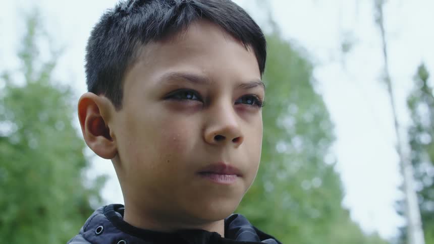 A boy wearing a dark colored jacket is standing outside with trees behind him