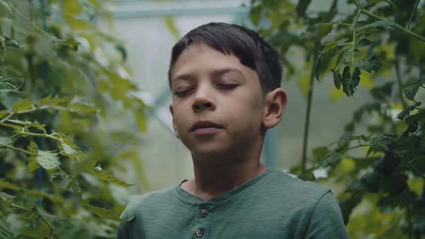 A boy is standing in a greenhouse and he closes his eyes and takes a deep breath
