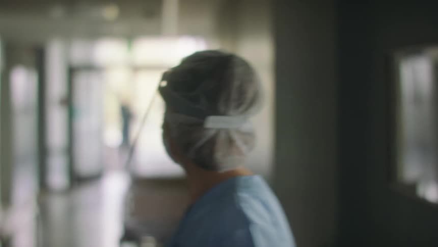 Female nurse stands staring in hospital corridor before turning and moving away rapidly
