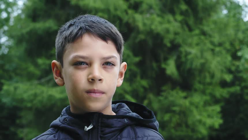 A boy wearing a dark colored jacket is standing outside with trees behind him
