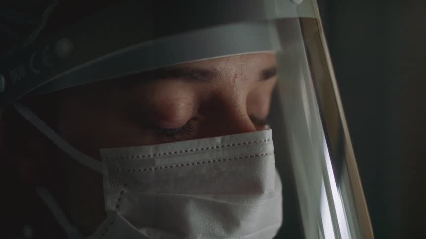 Medical personnel stands staring as she wears a medical mask and a face shield