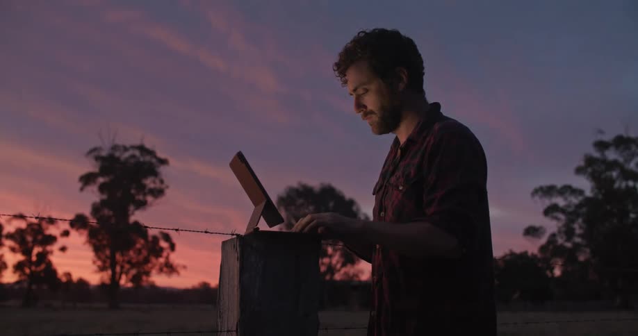 A man is outside typing on a laptop computer near a barbed wire fence