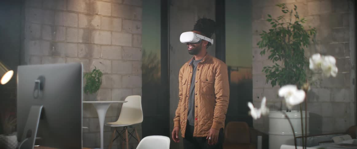 A man stands in front of a computer at home with a virtual reality headset on and looks around
