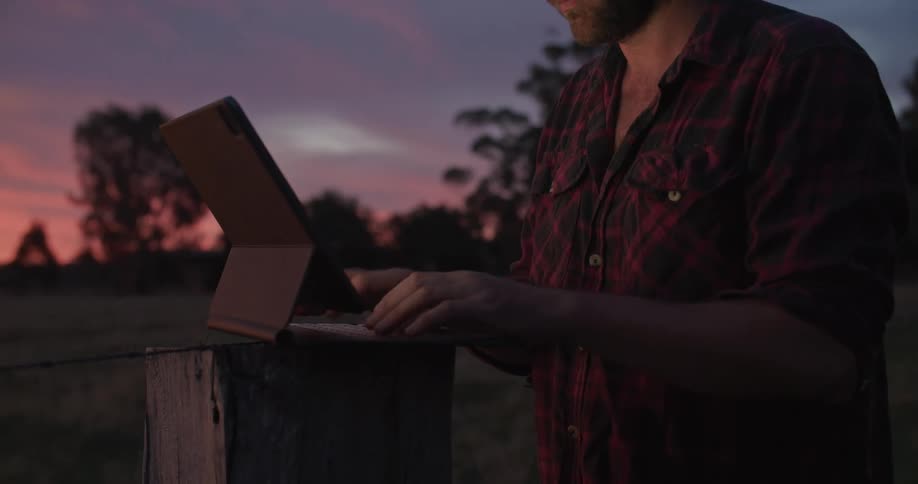 A man stands on a grassy field and types on a table as the sun sets