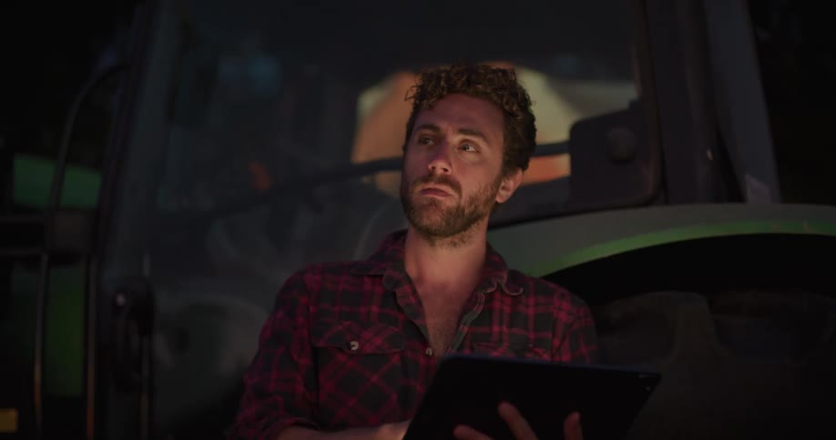 A man leans against a truck and types on a tablet and looks off in to the distance