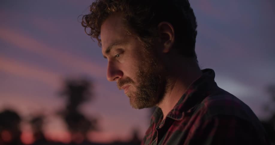 A man standing in a field types on a laptop as the sun sets