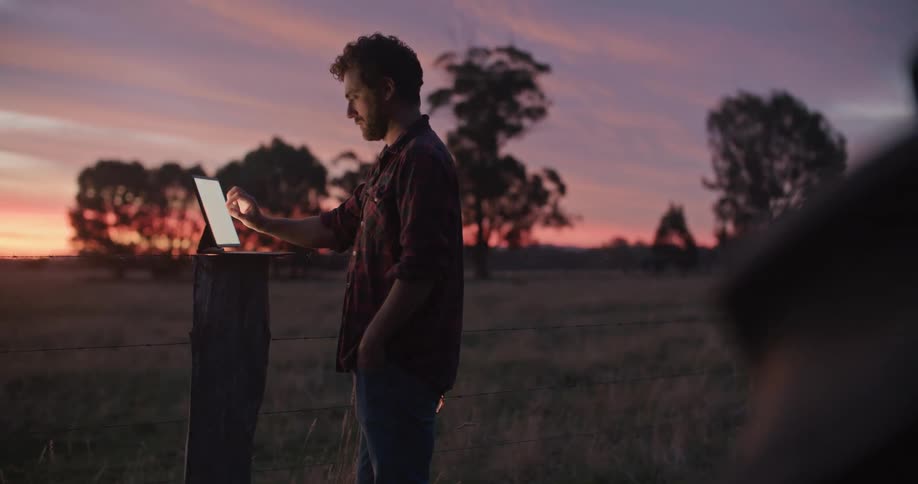 A man stands in a field and uses a laptop computer and then looks across the land