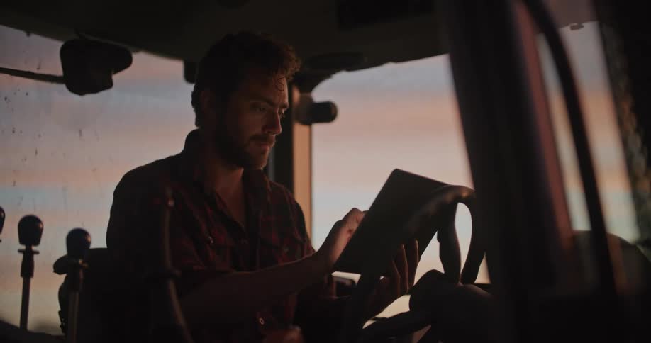 A young farmer is sitting in a tractor and looking at a tablet