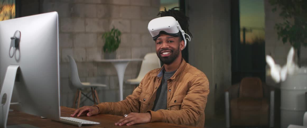 A man is sitting at a computer desk and wearing virtual reality gear
