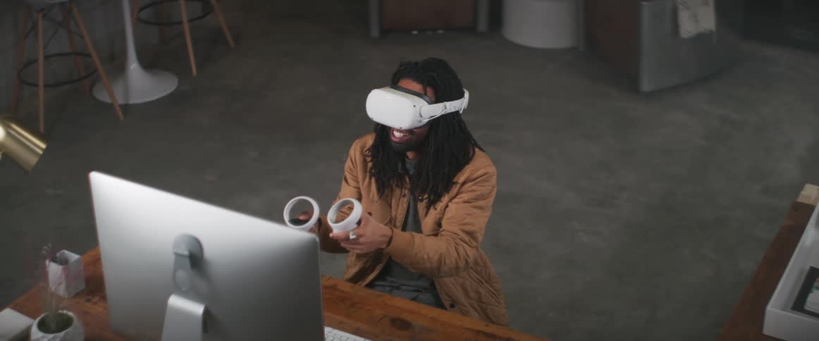 A man sits in front of a computer and plays a virtual reality game with goggles on